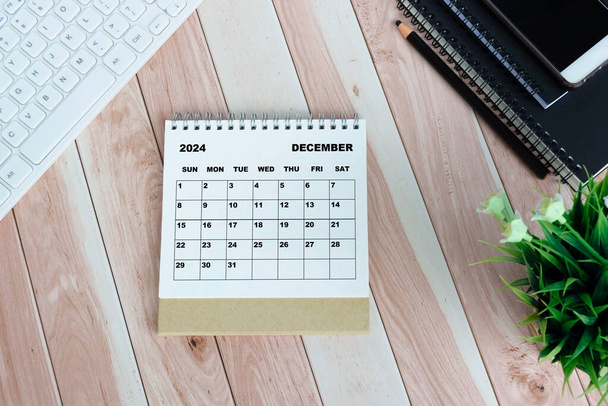 Bílý kalendář prosinec 2024 na dřevěném stole s klávesnicí, poznámkovými bloky, tužkou, květináčem a smartphonem. - Fotografie, Obrázek