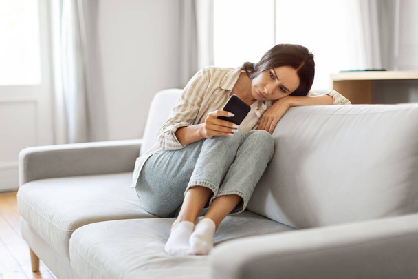 Αναστατωμένη νεαρή γυναίκα κοιτάζοντας οθόνη Smartphone, κάθεται στον καναπέ στο σπίτι, λυπημένος Millennial Lady περιμένοντας για σημαντική κλήση ή sms, Υποφέροντας εποχιακή κατάθλιψη, αισθάνεται μόνος και πληγωμένος - Φωτογραφία, εικόνα