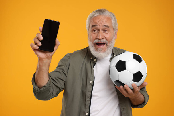 Happy senior sports fan with soccer ball showing smartphone on yellow background - Zdjęcie, obraz