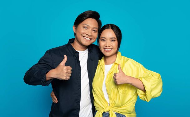 Positive young japanese guy and woman in casual show thumb up sign with hand, isolated on blue studio background. Emotions recommendation, approve gesture, good news sign - Foto, immagini