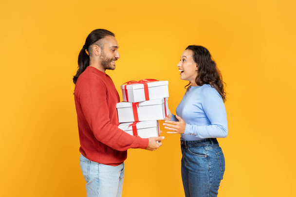 Happy young couple giving gifts to each other, yellow studio background, free space. Loving handsome man giving present to his excited wife or girlfriend. Valentines day celebration, profile side view - Photo, Image