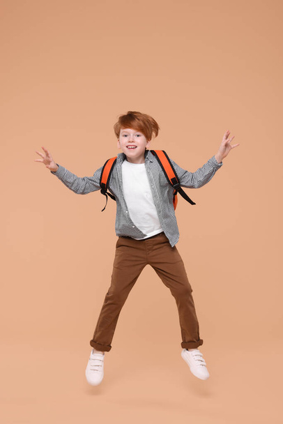 Cute schoolboy with backpack jumping on beige background - Фото, изображение