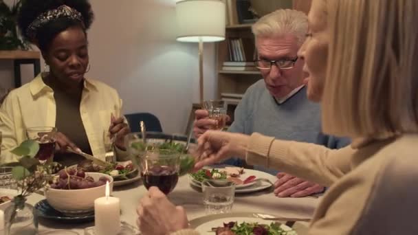 Над пліч-о-пліч кавказька жінка кладе салат на тарілки багатоетнічних членів сім'ї, сидячи за обіднім столом під час святкування - Кадри, відео