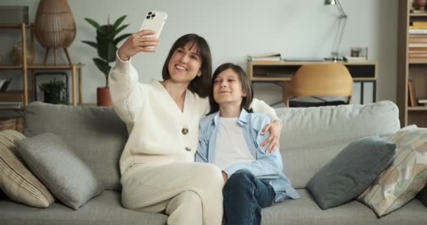 Mutter und Sohn halten einen besonderen Moment mit einem fröhlichen Selfie auf der Wohnzimmercouch fest. Ihr gemeinsames Lächeln strahlt Glück und Liebe aus und verkörpert eine herzerwärmende Szene familiärer Verbundenheit.. - Filmmaterial, Video