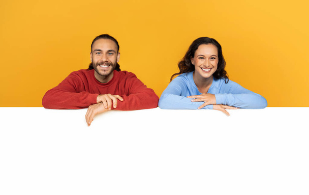 Positive cheerful young man and woman leaning on huge blank horizontal advertising board for your text, mockup, copy space, yellow studio background. Couple showing nice deal, great offer - Photo, Image