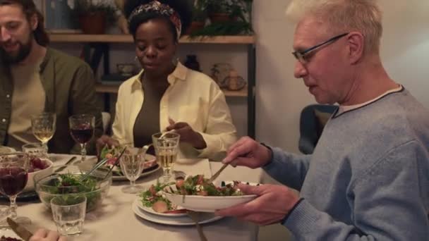Вид збоку дорослого чоловіка, який кладе салат з зеленню на тарілку, одночасно влаштовуючи урочисту вечерю з членами багатоетнічної сім'ї - Кадри, відео