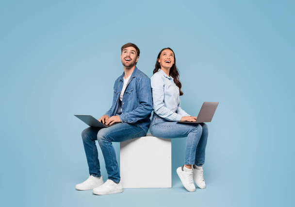 Giovani sposi europei seduti schiena a schiena, profondamente assorti nei loro computer portatili e guardando verso l'alto lo spazio libero su un rilassante sfondo blu studio, banner - Foto, immagini