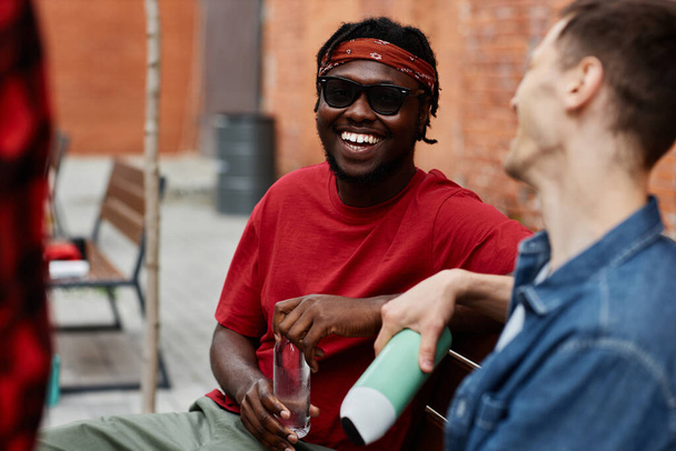 Bandana takan genç siyahi bir adamın portresi ve dışarıda arkadaşlarıyla sohbet ederken mutlu bir şekilde gülmesi. - Fotoğraf, Görsel