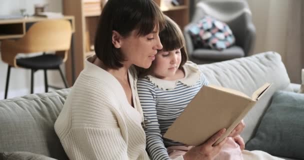 Mother and daughter share a moment on the living room couch, immersed in a book. Their shared love for reading creates a heartwarming scene of bonding and intellectual exploration within the family. - Footage, Video