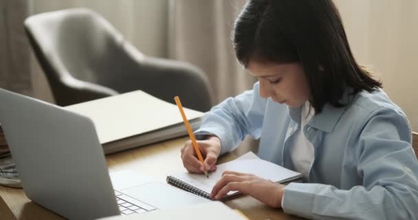 Mladý chlapec se věnuje soustředěnému studiu pomocí notebooku i notebooku. Jeho odhodlání je evidentní, protože bezproblémově kombinuje digitální zdroje s tradičními poznámkami. - Záběry, video