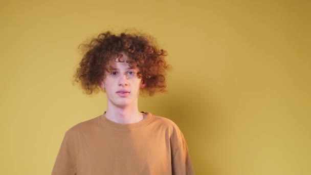 A guy with curly hair, wearing a t-shirt, looks at the camera with big eyes, raises his hands up, isolated on a yellow background in the studio. - Footage, Video