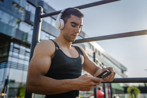 Egy férfi kaukázusi fiatal férfi stand a szabadban nyitott edzőteremben tornaterem használata mobiltelefon okostelefon fejhallgató üzeneteket küldeni SMS-t, vagy böngészni internet online alkalmazás képzés valós személy másolás hely - Fotó, kép