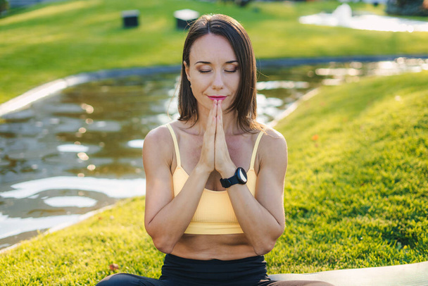 Caucasian slim fit woman sitting in lotus yoga pose on fitness mat in sportwear with hands in prayer position, in summer park. Front view. - Photo, Image