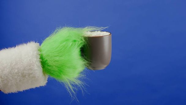 Grinchs green haired hand holds a cup of cocoa with marshmallows on isolated blue background. Gift kidnapper cosplay. Christmas and New Year celebration concept. Blue screen, chroma key - Foto, imagen