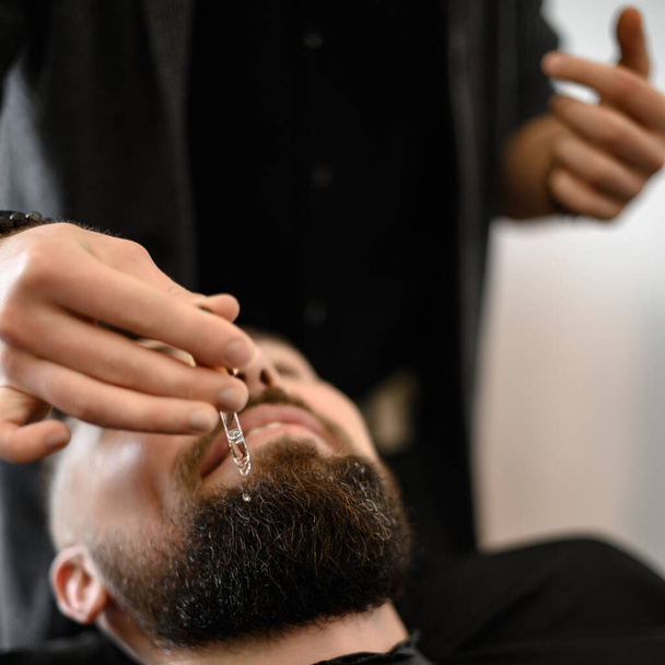 Парикмахер стилист наносит капли масла на клиентов бороду, чтобы увлажнить и смягчить. Уход за бородой. - Фото, изображение