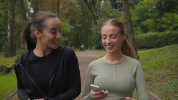 Zwei fröhliche athletische kaukasische Europäerinnen zwei Sportmädchen, die mit ihrem Handy im Stadtpark spazieren gehen, lächelnd aktive, fitte Sportlerinnen Läuferinnen unterhalten sich über hohe fünf gute Teamarbeit weibliche Freundschaft - Filmmaterial, Video