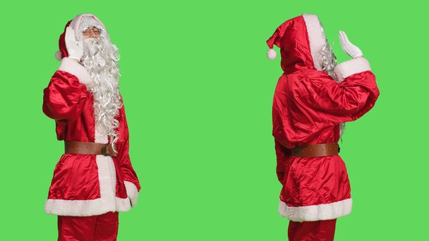 Людина в костюмі Санта-Клауса привітається на камеру, діючи радісно і впевнено на зеленому тлі. Святковий сезонний персонаж привітання людей або прощання, ізольований шаблон. - Фото, зображення
