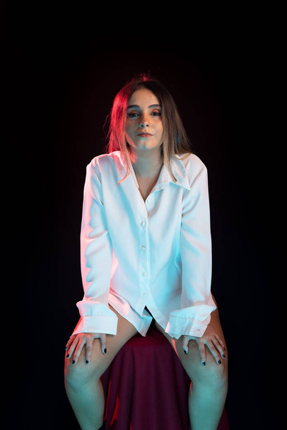 Beautiful, young attractive Caucasian woman sitting on a red stool wearing white outfit. Portrait in studio with black background. - Photo, Image