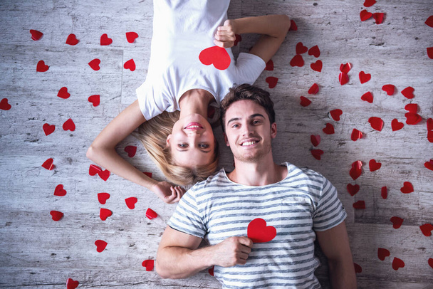 Вид зверху красива молода пара тримає червоні паперові серця, дивлячись на камеру і посміхаючись, лежачи на підлозі
 - Фото, зображення