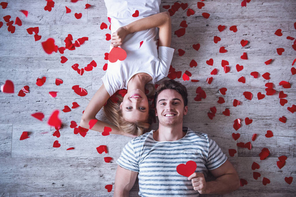 Vista superior de la hermosa pareja joven sosteniendo corazones de papel rojo, mirando a la cámara y sonriendo mientras está acostado en el suelo
 - Foto, imagen