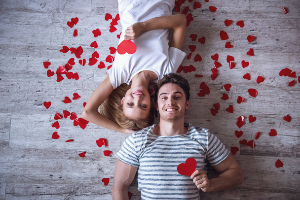 Top view of beautiful young couple holding red paper hearts, looking at camera and smiling while lying on the floor - Photo, Image