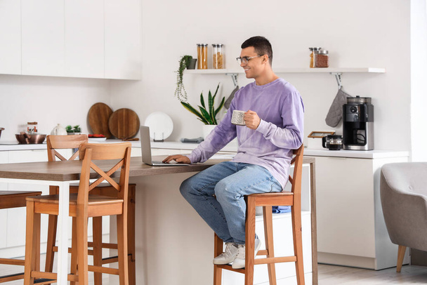 Schöner junger Mann mit einer Tasse Kaffee mit Laptop, während er zu Hause in der Küche sitzt - Foto, Bild