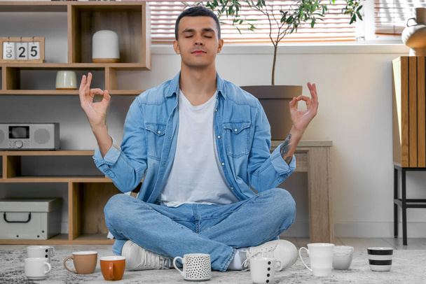 Handsome young man with many cups of coffee meditating on floor in living room - Photo, Image