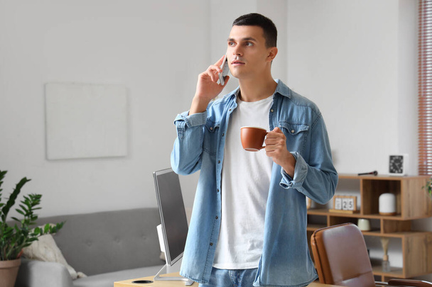自宅で携帯電話で話すコーヒーのカップを持つハンサムな若者 - 写真・画像