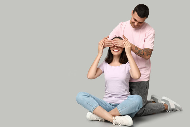 Young man covering eyes of his girlfriend while sitting on floor against grey background - Photo, Image