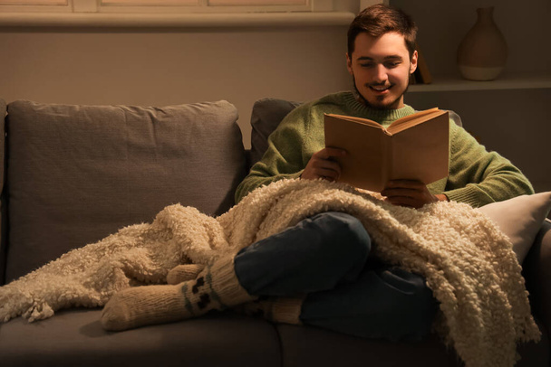 夜に自宅でソファーで本を読んでいる若者 - 写真・画像