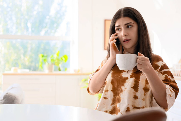 Νεαρή γυναίκα με φλιτζάνι καφέ κουτσομπολεύει από κινητό τηλέφωνο στην κουζίνα - Φωτογραφία, εικόνα