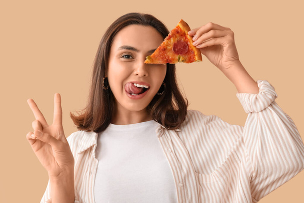Jeune femme tenant morceau de pizza savoureuse et montrant geste de victoire sur fond beige - Photo, image