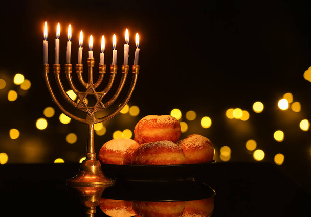 Menorah com velas acesas e donuts para a celebração Hanukkah na mesa escura contra luzes turvas - Foto, Imagem