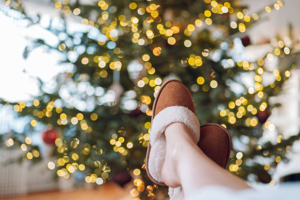 Рождественские праздники. Ноги в коричневых замшевых меховых тапочках на елке в гирляндах сзади. Зимний комфорт и праздники. Праздничное уютное настроение. - Фото, изображение