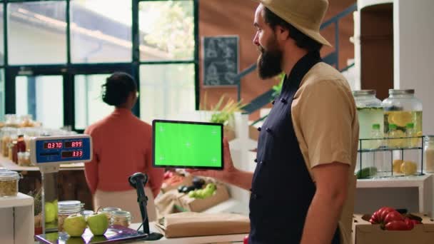 Storekeeper sostiene la tableta con pantalla verde en el supermercado ecológico de cero residuos, utilizando la maqueta de espacio de copia en el gadget móvil de pie en el mostrador de pago. Propietario de pequeña empresa presenta pantalla aislada. - Imágenes, Vídeo