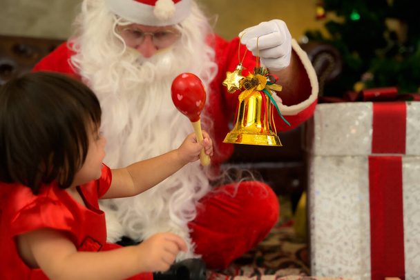 Маленькая девочка, сидящая вместе с Сантой Клаусом на полу и играющая с игрушечным колокольчиком дома. Празднование Рождества и Дня благодарения. - Фото, изображение