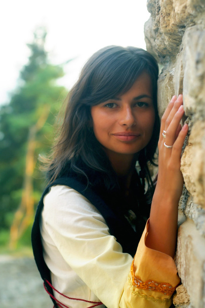 Une belle jeune femme avec de longs cheveux foncés et une robe historique à un mur de pierre ancienne rugueuse
 - Photo, image