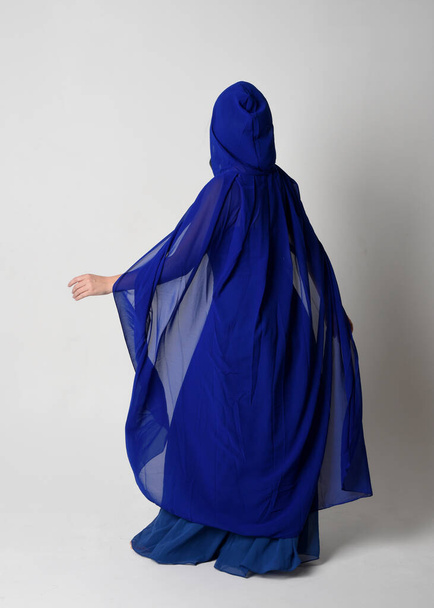 Retrato de cuerpo entero de hermosa joven modelo femenino elegante vestido de bola de fantasía azul y capa que fluye con capucha. Posar de pie, alejándose mirando hacia atrás. Aislado en el fondo del estudio. - Foto, Imagen