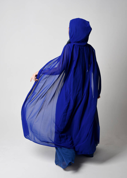 Πλήρες μήκος πορτρέτο της όμορφης νεαρής γυναίκας μοντέλο κομψό μπλε φόρεμα μπάλα φαντασίας και ρέουσα κάπα με κουκούλα. Στέκομαι όρθιος, περπατώντας προς τα πίσω. Απομονωμένο στο φόντο του στούντιο Γουίτ. - Φωτογραφία, εικόνα