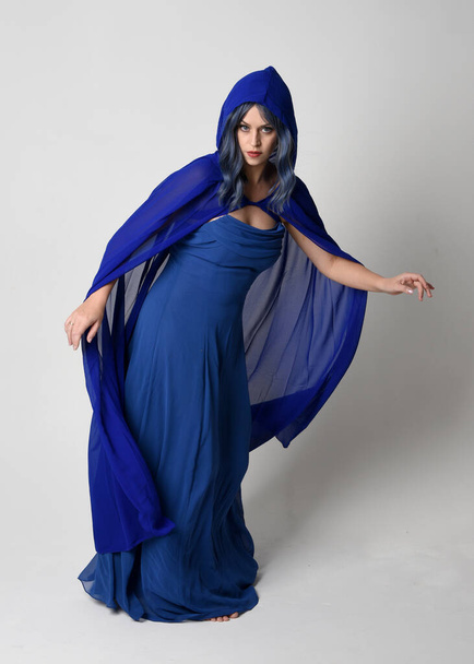 Retrato de cuerpo entero de una hermosa modelo femenina que viste un elegante vestido de fantasía azul y una capa fluida con capucha. Posada de pie, con los brazos gestuales extendidos. Aislado sobre fondo blanco estudio. - Foto, Imagen