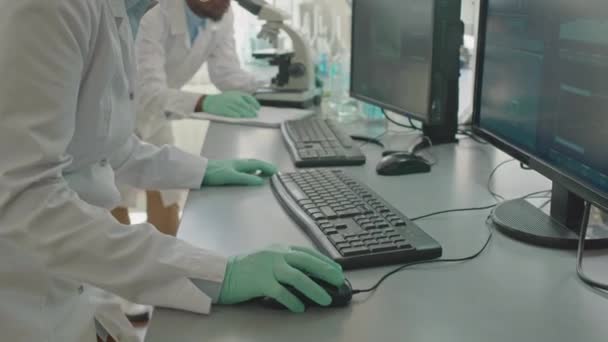 Inclinazione di scienziata caucasica in camice bianco guardando lo schermo del computer durante l'analisi della ricerca, lavorando con il collega afroamericano maschio in laboratorio - Filmati, video