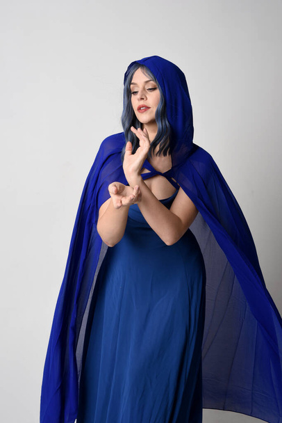Close up portrait of beautiful female model wearing elegant fantasy blue ball gown, flowing cape with hooded cloak.Gestural arms reaching out. Isolated on white studio background. - Foto, Imagem