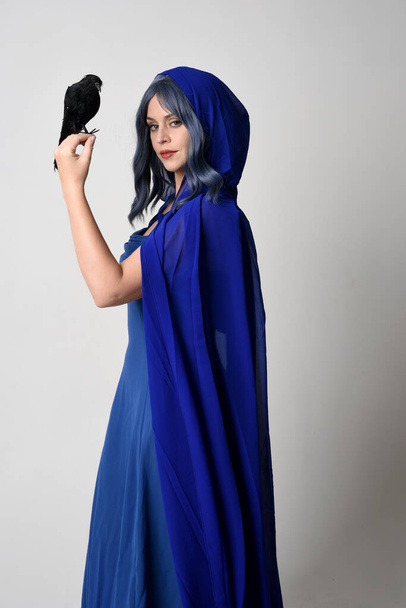 Ritratto ravvicinato di bella modella donna che indossa elegante fantasia abito da ballo blu, mantello fluente con cappuccio. Con in mano un finto uccello nero. Isolato su studio bianco  - Foto, immagini