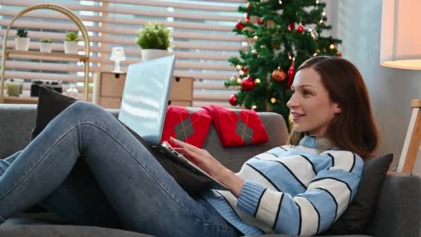 Mujer caucásica satisfecha en suéter caliente usando el ordenador portátil, relajarse en el sofá cerca del árbol de Navidad en las vacaciones de invierno. - Metraje, vídeo