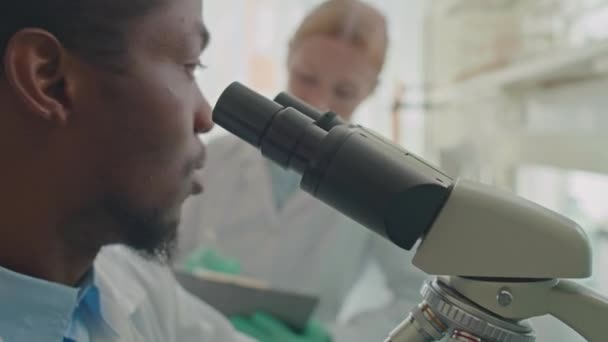 Strzał przechyłu profesjonalnego afroamerykańskiego mikrobiologa płci męskiej przy użyciu mikroskopu i dzielenie się wynikami z białą koleżanką, współpracownikami badań naukowych w laboratorium - Materiał filmowy, wideo