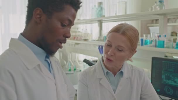 Abbassamento di due scienziati multietnici maschili e femminili in camici bianchi che guardano tablet digitale e discutono i risultati della ricerca mentre lavorano in un laboratorio modernamente attrezzato - Filmati, video