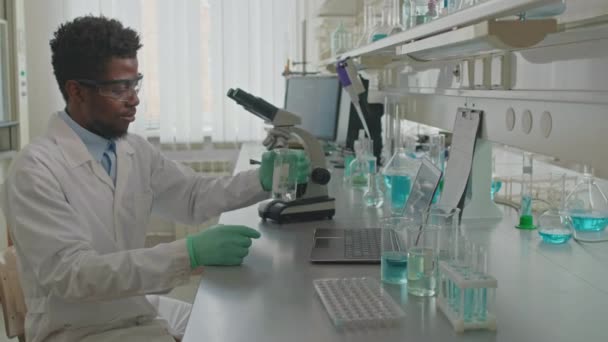 Colpo medio di giovane scienziato afroamericano di sesso maschile che utilizza tazza di misurazione di vetro mentre conduce esperimenti chimici in laboratorio e annota note sul computer portatile - Filmati, video
