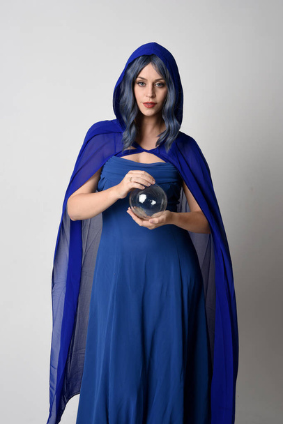 Close up portrait of beautiful female model wearing elegant fantasy blue ball gown, flowing cape with hooded cloak. Holding a crystal glass seer orb. Isolated on white studio background. - Photo, Image