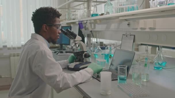 Середній знімок афроамериканського біохіміка чоловічої статі з використанням мікроскопа та проведення хімічних експериментів під час роботи за столом в лабораторії - Кадри, відео