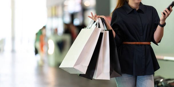 Γυναίκα χρησιμοποιώντας smartphone κρατώντας μαύρη τσάντα ψώνια Παρασκευή, ενώ στέκεται στο πλάι με το εμπορικό κέντρο. - Φωτογραφία, εικόνα